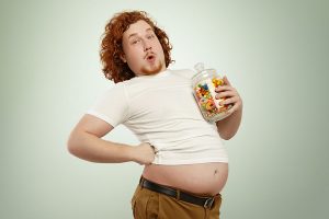 Happy overweight redhead man having joyful look, holding jar of goodies, keeping hand on waist, let his belly hang out of t-shirt. Obese fat male eating junk food, posing at blank studio wall